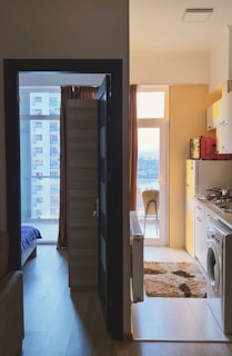 A small apartment space with two main areas visible. On the left, there's a partially open door leading to a room with a window showing a view of another building. The room has a bed and a couch. On the right, the kitchenette area includes a washing machine, stove with pots, cupboards, and a small table near the window with natural light coming through.