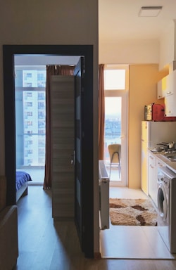 A small apartment space with two main areas visible. On the left, there's a partially open door leading to a room with a window showing a view of another building. The room has a bed and a couch. On the right, the kitchenette area includes a washing machine, stove with pots, cupboards, and a small table near the window with natural light coming through.