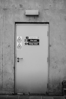 A grey metal door set into a concrete wall, featuring multiple warning signs and labels. The door has a handle and a lock, and it is marked as a fire exit with a clear 'Keep clear' sign. Two triangular warning signs indicate potential chemical hazards, emphasizing safety precautions.