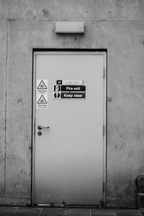 A grey metal door set into a concrete wall, featuring multiple warning signs and labels. The door has a handle and a lock, and it is marked as a fire exit with a clear 'Keep clear' sign. Two triangular warning signs indicate potential chemical hazards, emphasizing safety precautions.