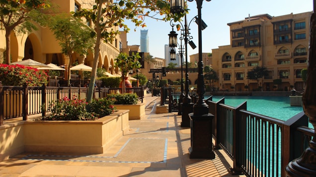 A sunny urban setting features a path lined with ornate street lamps and bordered by water on one side and Mediterranean-style buildings on the other. Trees and red flowers add greenery, while blue-tinted water reflects the clear sky. Several seating areas with umbrellas provide a shaded retreat.