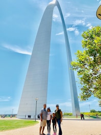 A tall, modern architectural arch set against a clear blue sky, with a well-maintained grassy area in the foreground. A family of four stands near the base of the structure, appearing to enjoy a sunny day outdoors. The arch is the focal point of the image, with foliage from a nearby tree visible on the right.
