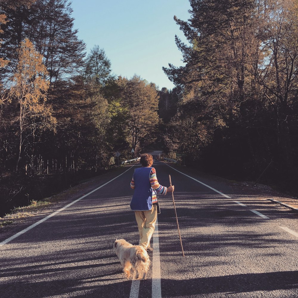 man in black jacket and brown pants walking with brown dog on road during daytime