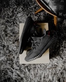A pair of black sneakers placed on a shoebox, resting on a textured gray carpet. The shoes have a modern design with a ribbed sole and a knitted upper. There's also a partial view of a wooden leg of a chair with a patterned seat in the upper right corner.
