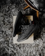 A pair of black sneakers placed on a shoebox, resting on a textured gray carpet. The shoes have a modern design with a ribbed sole and a knitted upper. There's also a partial view of a wooden leg of a chair with a patterned seat in the upper right corner.