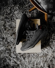 A pair of black sneakers placed on a shoebox, resting on a textured gray carpet. The shoes have a modern design with a ribbed sole and a knitted upper. There's also a partial view of a wooden leg of a chair with a patterned seat in the upper right corner.