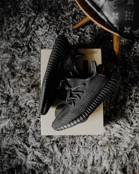 A pair of black sneakers placed on a shoebox, resting on a textured gray carpet. The shoes have a modern design with a ribbed sole and a knitted upper. There's also a partial view of a wooden leg of a chair with a patterned seat in the upper right corner.