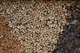 A neatly stacked pile of split firewood featuring a variety of wood types, some with lighter tan colors and others darker, possibly charred. The logs are cut into small, consistent pieces, filling the entire frame with an orderly pattern.