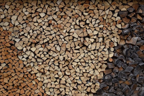 A neatly stacked pile of split firewood featuring a variety of wood types, some with lighter tan colors and others darker, possibly charred. The logs are cut into small, consistent pieces, filling the entire frame with an orderly pattern.