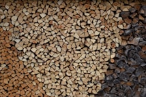 A neatly stacked pile of split firewood featuring a variety of wood types, some with lighter tan colors and others darker, possibly charred. The logs are cut into small, consistent pieces, filling the entire frame with an orderly pattern.