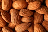 A close-up view of a heap of almonds. The nuts display a textured, brown surface and are arranged closely together, filling the frame entirely.