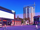 A modern commercial area with a multistory building under construction in the background. The foreground features a small plaza with people walking and pigeons scattered around. On the left, a large building with bold signage and glass storefront windows is visible, and a smaller semi-circular building on the right. A yellow construction crane is located near the tall building under construction.