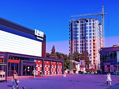A modern commercial area with a multistory building under construction in the background. The foreground features a small plaza with people walking and pigeons scattered around. On the left, a large building with bold signage and glass storefront windows is visible, and a smaller semi-circular building on the right. A yellow construction crane is located near the tall building under construction.