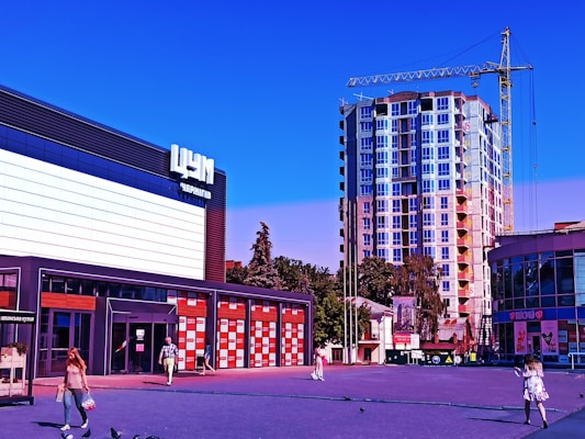 A modern commercial area with a multistory building under construction in the background. The foreground features a small plaza with people walking and pigeons scattered around. On the left, a large building with bold signage and glass storefront windows is visible, and a smaller semi-circular building on the right. A yellow construction crane is located near the tall building under construction.