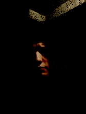 Close-up of the mysterious grey man’s face half-hidden in shadow, with a textured grey background.