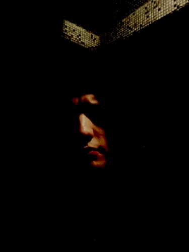 A shadowy face is partially illuminated, creating a mysterious and intense atmosphere. The background is mostly dark, with a hint of texture from a mosaic pattern in the upper area.