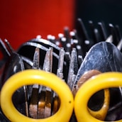 A collection of kitchen utensils featuring forks, knives, spoons, and scissors with yellow handles is arranged in a disorganized manner. The metal surfaces of the utensils create a metallic texture, and the focus is on the contrast between the shiny surfaces and the vibrant yellow color of the scissors' handles.