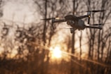 A drone is captured in flight against the backdrop of a setting sun. The sun casts a warm, golden light across the scene, highlighting the silhouette of barren trees in the background. The drone's propellers are visibly spinning, and it appears to be a modern, sleek model.
