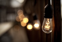 A soft-focus image highlighting a string of illuminated vintage-style light bulbs, with a distinct large bulb in the foreground. The warm glow contrasts with a blurred, dark background creating a cozy, inviting atmosphere.