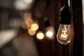 A soft-focus image highlighting a string of illuminated vintage-style light bulbs, with a distinct large bulb in the foreground. The warm glow contrasts with a blurred, dark background creating a cozy, inviting atmosphere.