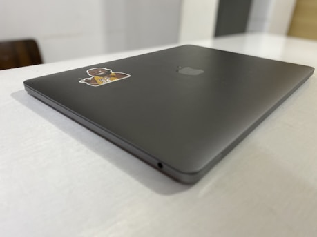A sleek, closed laptop with a matte finish is positioned at an angle on a light-colored table. The laptop has an Apple logo on the lid and a sticker featuring a cartoon character in a sports jersey.