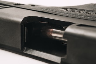 A close-up view of a firearm with a visible bullet in the chamber. The metallic surface of the bullet contrasts with the dark, textured material of the gun. The angle provides a detailed look at the mechanics of the loading area.