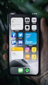 A hand holding a smartphone displaying its home screen filled with various app icons, including social media, photography, and utility apps. The background shows a few leaves, adding a natural element to the scene. The time is shown as 5:08, and the date is set to Monday the 6th. Widgets present include a weather forecast for Ottawa, currently showing a temperature of 31°C with mostly sunny conditions.