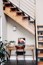 A wooden staircase with metal railings runs diagonally across the upper portion of a minimalist workspace. Below the staircase, a wooden desk is neatly organized with a small plant, a closed laptop displaying abstract art, and a keyboard, accompanied by a chair. The interior has neutral-colored walls and a few green plants adding a touch of nature.