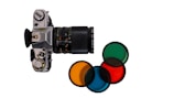 A vintage camera with a long zoom lens, positioned vertically against a plain white background. Alongside the camera, four lens filters are neatly arranged, each exhibiting different colors including green, orange, red, and blue. The camera appears well-crafted with a mechanical dial and a leather grip.