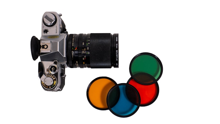 A vintage camera with a long zoom lens, positioned vertically against a plain white background. Alongside the camera, four lens filters are neatly arranged, each exhibiting different colors including green, orange, red, and blue. The camera appears well-crafted with a mechanical dial and a leather grip.