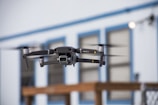 A drone is captured in mid-flight with its rotors in motion, set against a blurred backdrop of a building with blue trim and paneled windows. The drone appears to be technologically advanced and well-designed, indicating its suitability for aerial photography or videography.