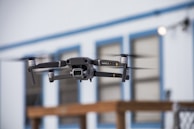 A drone is captured in mid-flight with its rotors in motion, set against a blurred backdrop of a building with blue trim and paneled windows. The drone appears to be technologically advanced and well-designed, indicating its suitability for aerial photography or videography.