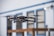 A drone is captured in mid-flight with its rotors in motion, set against a blurred backdrop of a building with blue trim and paneled windows. The drone appears to be technologically advanced and well-designed, indicating its suitability for aerial photography or videography.
