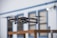 A drone is captured in mid-flight with its rotors in motion, set against a blurred backdrop of a building with blue trim and paneled windows. The drone appears to be technologically advanced and well-designed, indicating its suitability for aerial photography or videography.