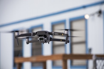 A drone is captured in mid-flight with its rotors in motion, set against a blurred backdrop of a building with blue trim and paneled windows. The drone appears to be technologically advanced and well-designed, indicating its suitability for aerial photography or videography.