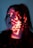 A person's face is illuminated by red light projections, possibly displaying characters or symbols. The person has closed eyes and dark hair, creating a contemplative or serene expression. The lighting casts dramatic shadows, enhancing the mysterious atmosphere.