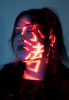 A person's face is illuminated by red light projections, possibly displaying characters or symbols. The person has closed eyes and dark hair, creating a contemplative or serene expression. The lighting casts dramatic shadows, enhancing the mysterious atmosphere.