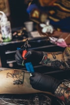 A tattoo artist is in the process of applying a detailed tattoo to a person's leg. The design features a horse, intricate patterns, and decorations. The artist is wearing black gloves and using a tattoo machine.