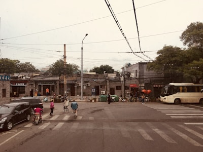 An urban street scene with several people riding bicycles and scooters past small shops and buildings. A large bus and a few cars are stopped at an intersection. Overhead, numerous electrical wires straddle the street, and trees are visible in the background.