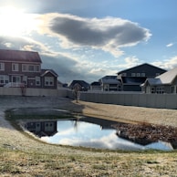 A suburban neighborhood scene featuring several modern houses with sloped roofs, surrounded by a white fence. A small pond in the foreground reflects the houses and sky. The grassy area around the pond appears slightly frosty, suggesting a cool or early winter setting. The sky is partly cloudy with sunlight peeking through, casting soft shadows.