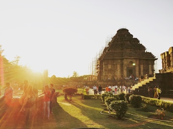 A large ancient stone temple is bathed in the warm glow of the setting sun, with visitors walking around the well-maintained gardens. People are scattered throughout the area, either exploring the site or sitting on stone ledges, enjoying the view.