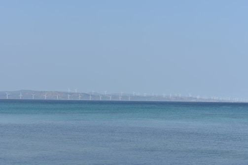 A vast expanse of ocean meets the horizon where numerous wind turbines are visible in the distance, set against a clear blue sky.