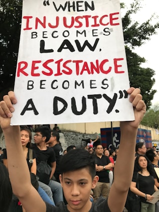 A young person holds a sign at a protest with the quote 'When injustice becomes law, resistance becomes a duty.' A group of people in the background are also participating in the protest, conveying a sense of solidarity. They stand in front of a wall with some graffiti and surrounded by trees.