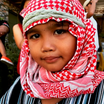 A young child wearing a red and white patterned headscarf and a striped shirt, looking towards the camera with a calm expression. The background includes a hint of adults engaged in conversation.