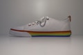 A white canvas sneaker is displayed against a plain background. The shoe features a rainbow-colored stripe running along the midsole, adding a vibrant touch to its design. It has white laces and multicolored eyelets that match the rainbow theme.