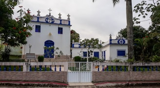 A small, white chapel with blue accents stands surrounded by green foliage. The structure features religious symbols on its roof, blue doors, and windows. A white fence with a gate encloses the area, which includes lamps and lush plants.