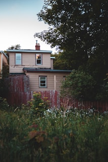 A two-story house with beige siding and a dark roof is surrounded by a lush garden. The structure is partially obscured by overgrown grass and shrubs. A weathered, red wooden fence borders the property, and there are tall trees in the background.