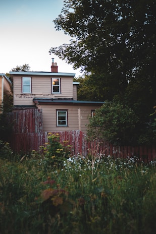 A two-story house with beige siding and a dark roof is surrounded by a lush garden. The structure is partially obscured by overgrown grass and shrubs. A weathered, red wooden fence borders the property, and there are tall trees in the background.