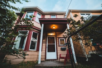 A two-story house with beige siding and red trim, featuring a front door with the number 55. The entrance is illuminated by a warm light above, and there are visible windows on both floors. The house is partly hidden by green foliage, and in front of the entrance, a red chair can be seen. A mesh metal fence is in the foreground.