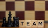Wooden blocks with the word 'TEAM' are placed next to black chess pieces, including pawns and a king. The background features a blurred chessboard pattern with alternating light and dark squares.